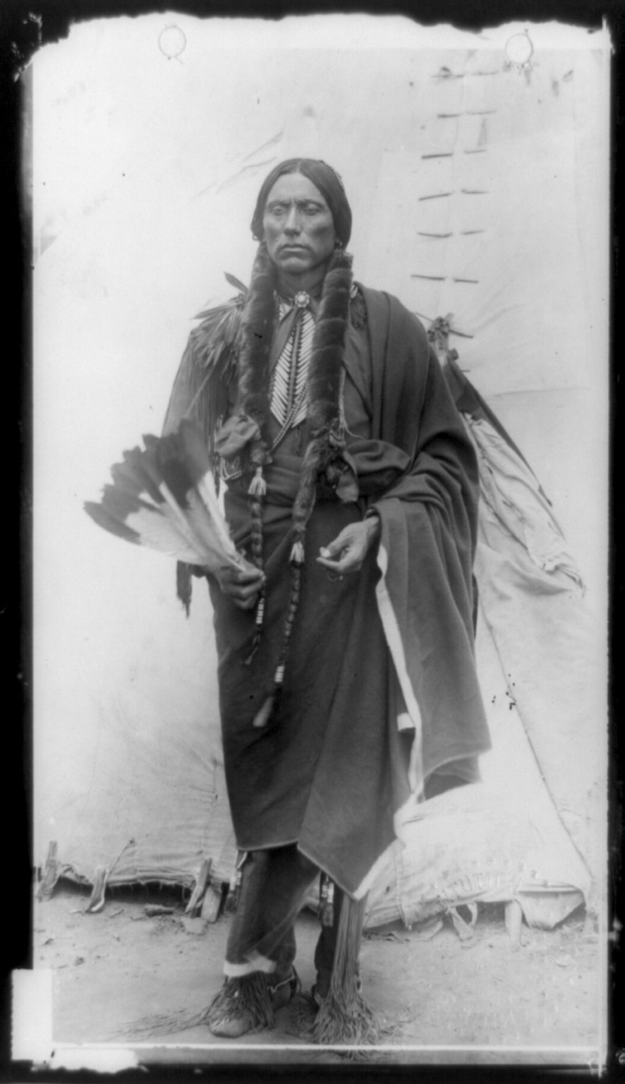 Quanah Parker, Comanche chief, full-length portrait holding feathers in front of a tepee